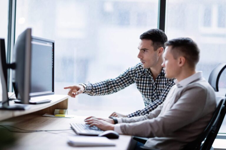 Two men in a discussion, with one pointing at the computer monitor, both focused on the screen, indicating a collaborative work environment